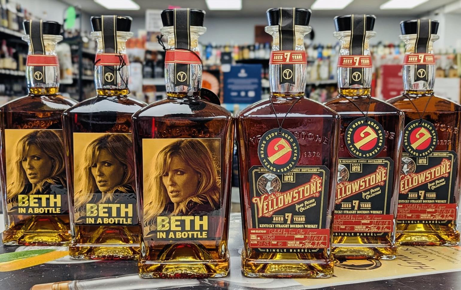 Row of bourbon bottles on a store counter, including three “Beth in a Bottle” labels and three Yellowstone Aged 7 Years Kentucky straight bourbon whiskey bottles, with liquor shelves blurred in the background.