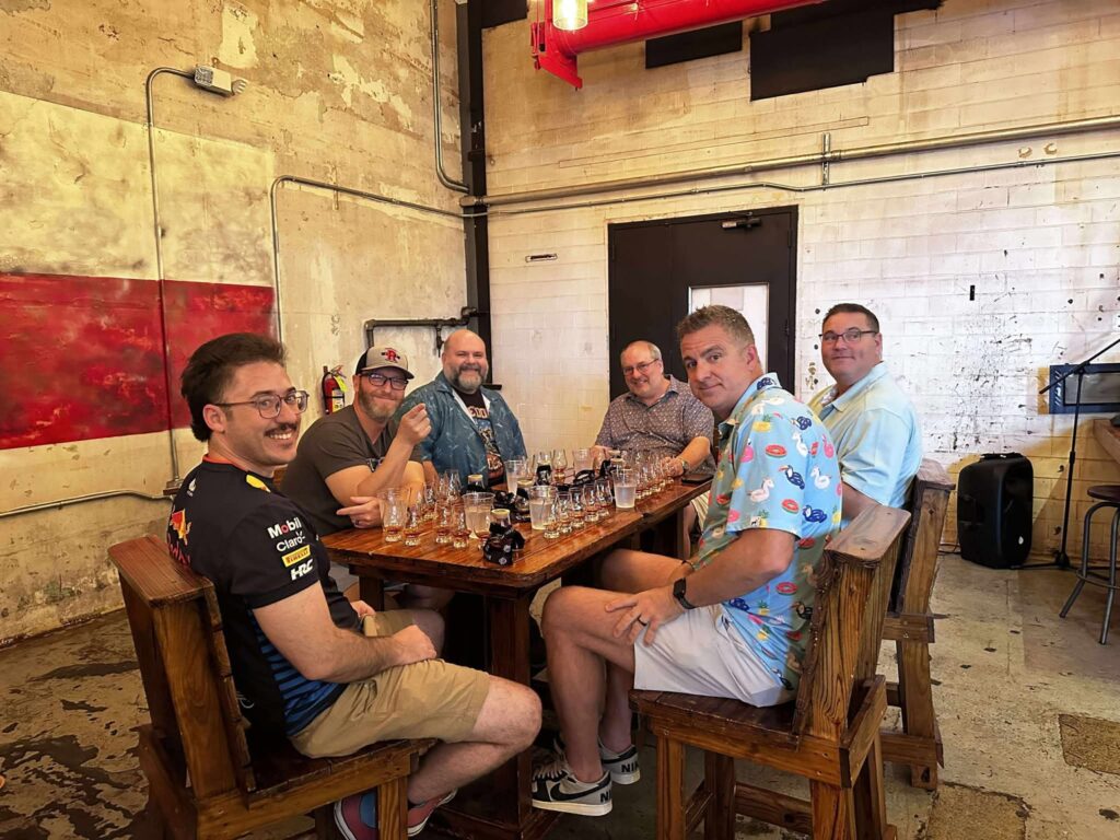 Six men seated at a rustic wooden table in an industrial taproom, smiling toward the camera with tasting glasses and beer bottles spread across the table.