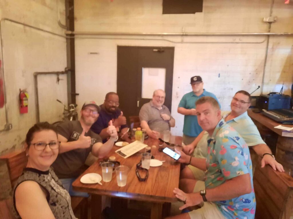 Seven adults gathered around a wooden table in a casual indoor space, smiling at the camera with drinks, plates, and a phone on the table.
