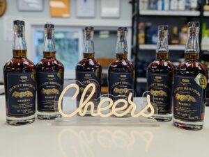 Six bottles of Elliott Brothers Reserve bourbon lined up on a bar counter behind a glowing “Cheers” sign.