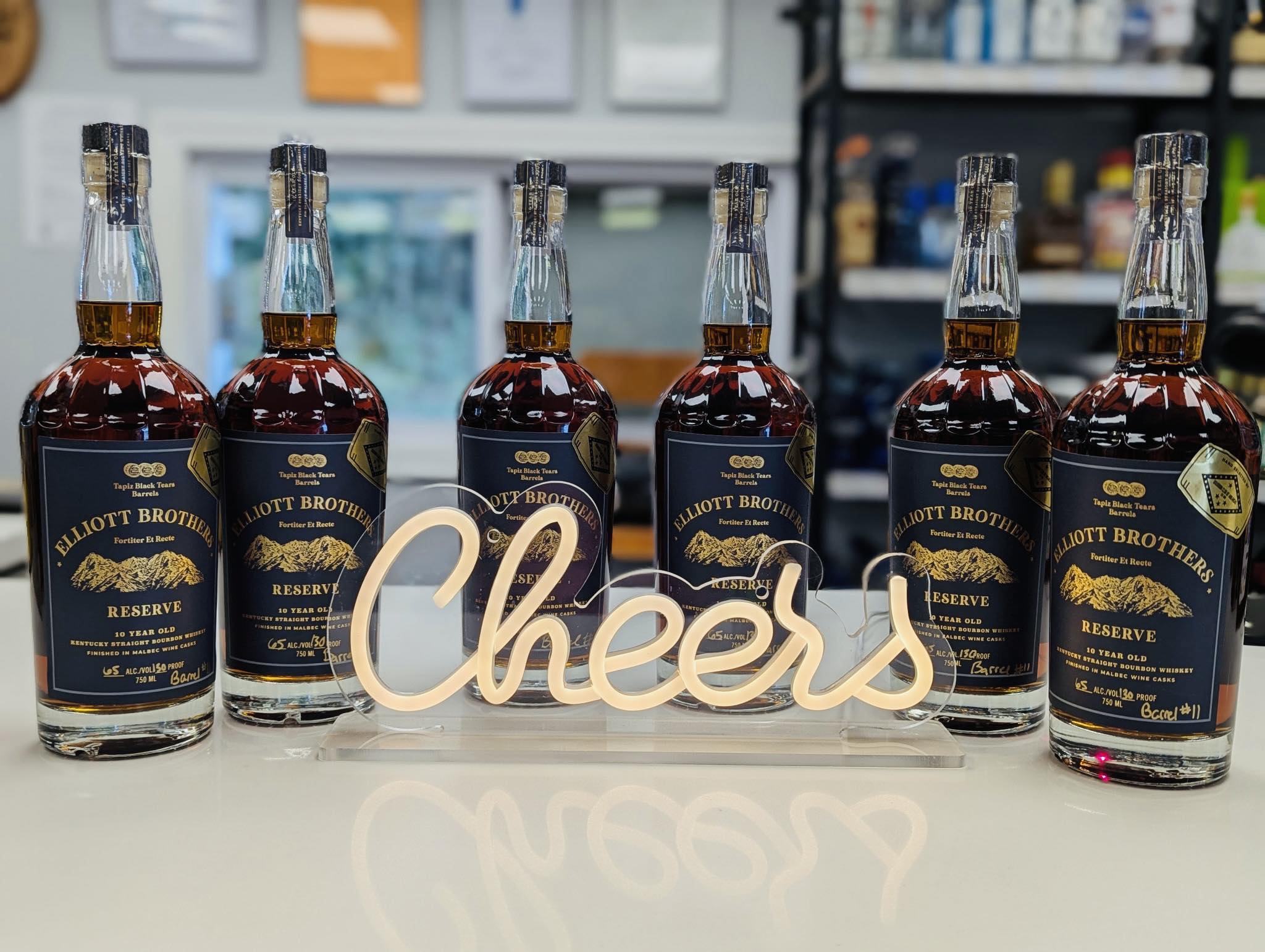 Six bottles of Elliott Brothers Reserve bourbon lined up on a bar counter behind a glowing “Cheers” sign.