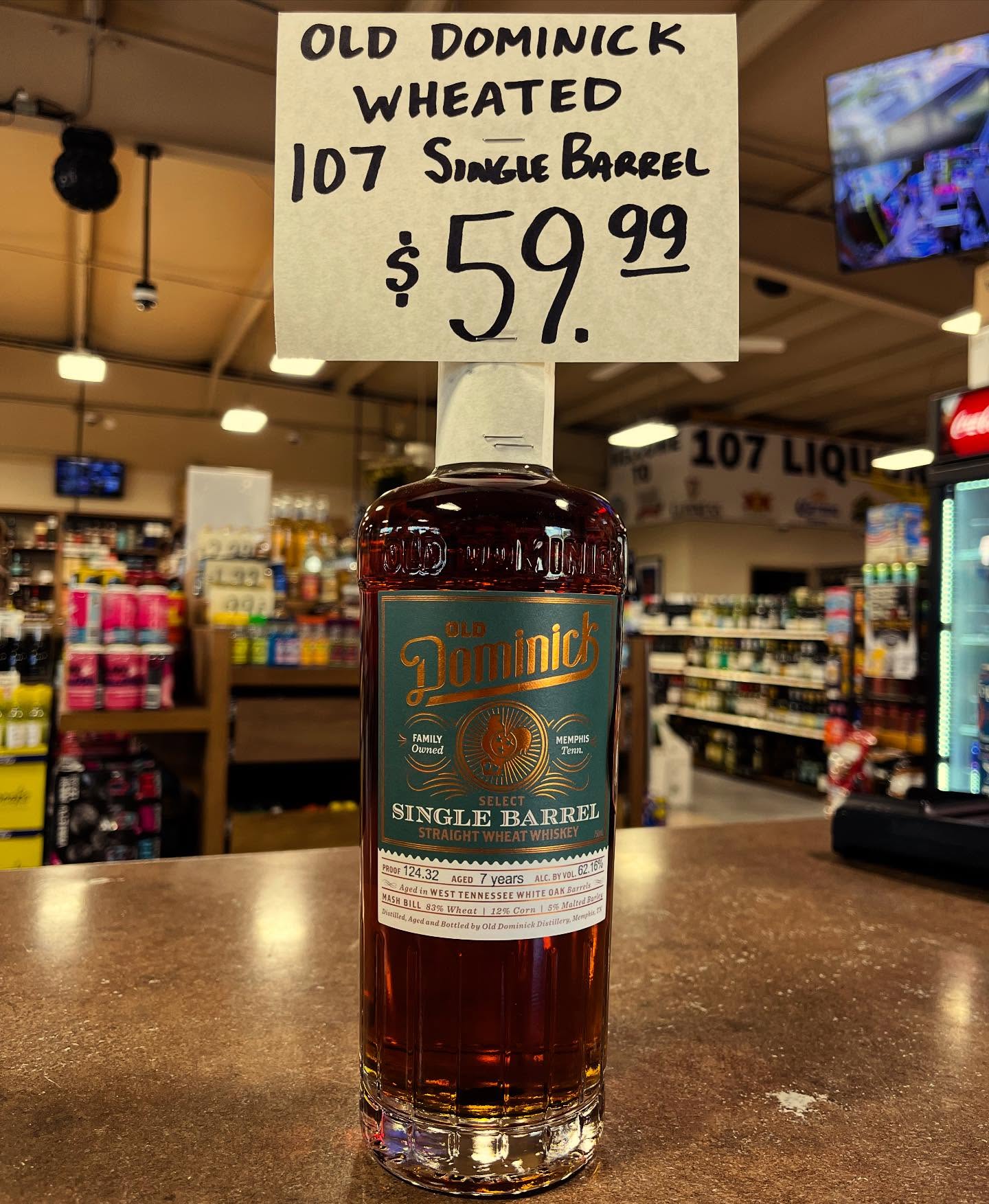 Old Dominick Wheated 107 Single Barrel straight wheat whiskey bottle on a liquor store counter with a handwritten sign above showing the price $59.99.