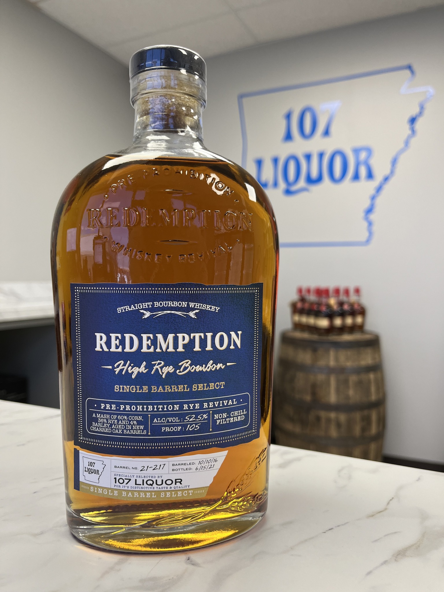 A close-up of a bottle of Redemption High Rye Bourbon, featuring a label that says 'Redemption High Rye Bourbon Single Barrel Select' with details of its distillation, bottling date, and proof. In the background, a sign for '107 Liquor' is visible along with whiskey barrels.