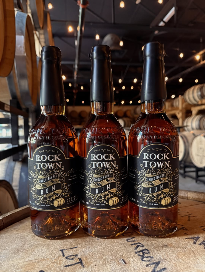A close-up of three bottles of Rock Town Single Barrel Rum arranged in a row on top of a wooden barrel. The bottles contain amber liquid and feature black wax seals and ornate black labels with gold lettering. The glass bottles are embossed with 'Rock Town Distillery.' The background is a soft-focus view of a barrel warehouse with stacked casks and warm string lights.