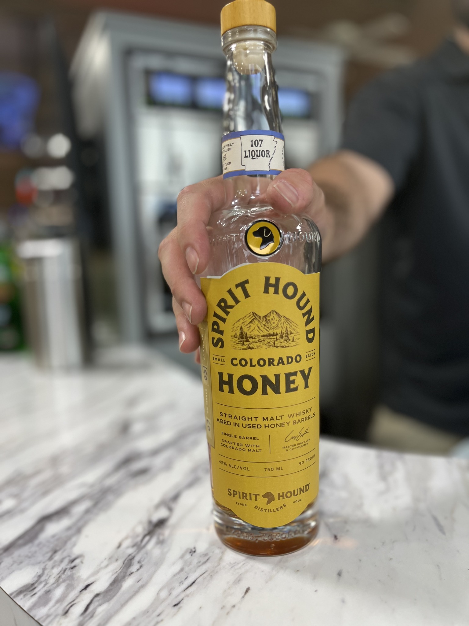 Hand holding a bottle of Spirit Hound Colorado Honey straight malt whisky on a marble countertop, with a blurred bar background.