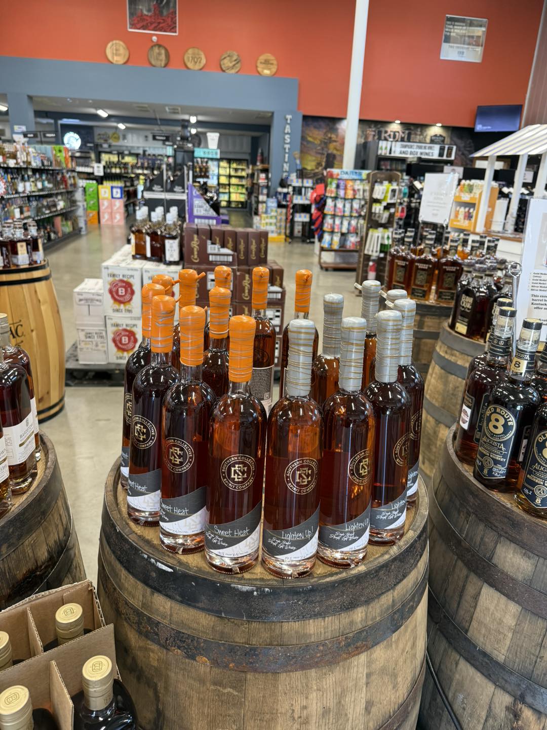 Display of Southern Collective Spirits bottles on a wooden barrel in a liquor store aisle, with shelves of spirits and a tasting area in the background.