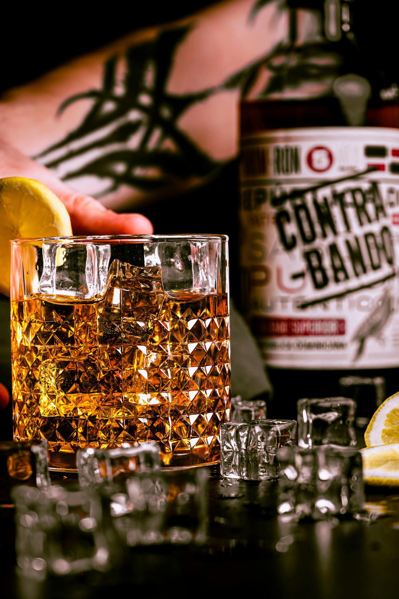 Close-up of a whisky glass with ice and lemon, set in a bar with a tattooed hand.