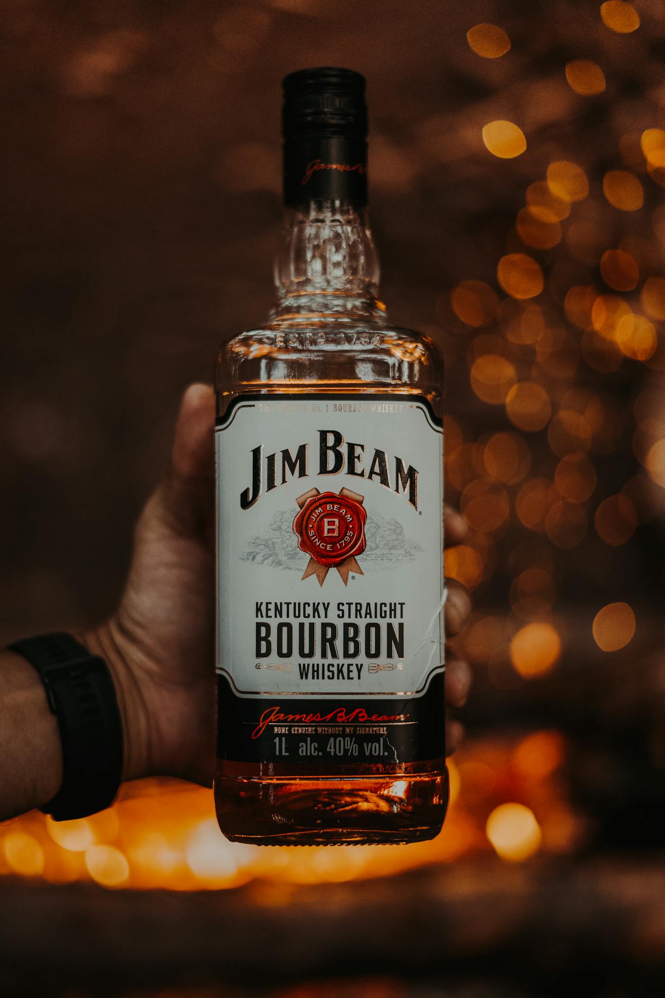Warm-toned image of a hand holding a Jim Beam Bourbon bottle with bokeh background.