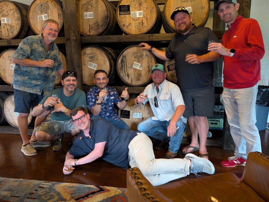 Seven men pose in front of stacked wooden bourbon barrels, smiling and holding tasting glasses; one man lies playfully on the floor in the foreground.