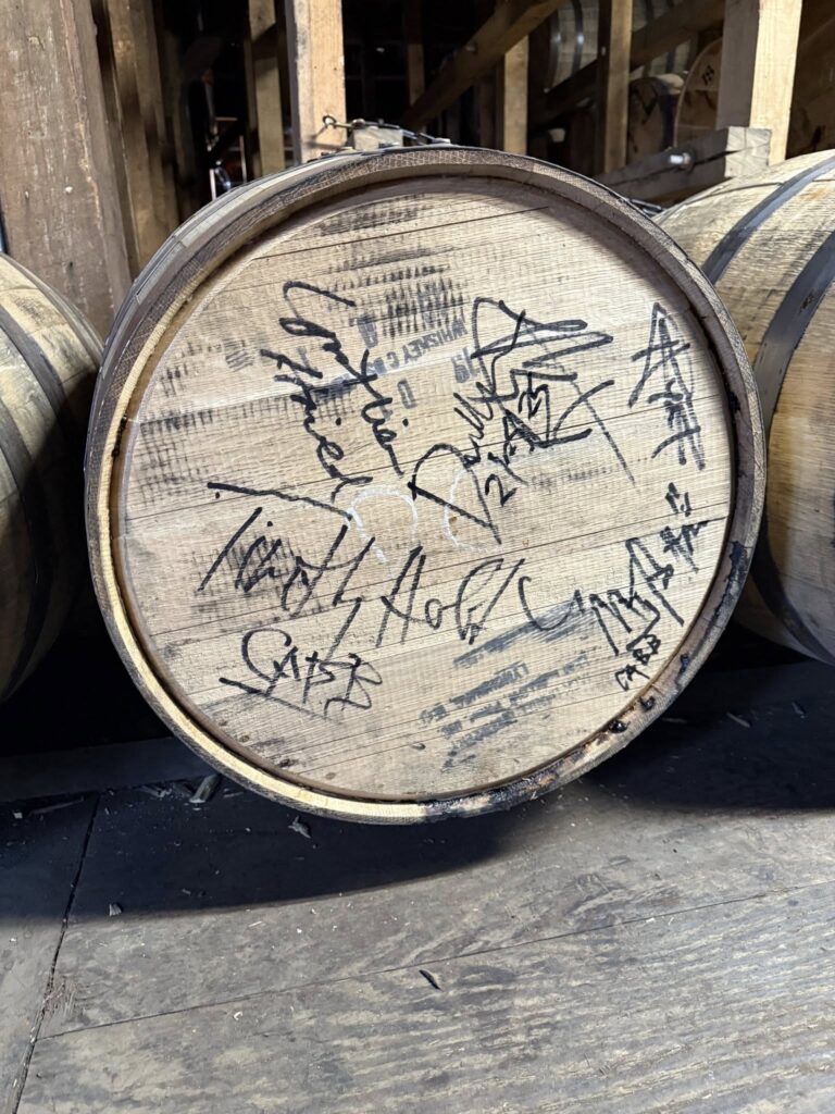 Close-up of a wooden whiskey barrel end with black marker signatures and graffiti, resting on a distillery warehouse floor among other barrels.