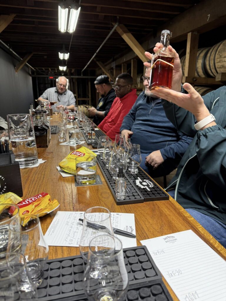 Group whiskey tasting in a barrel warehouse, with guests seated at a long table of tasting glasses and notes while one person holds up a bottle of amber whiskey.