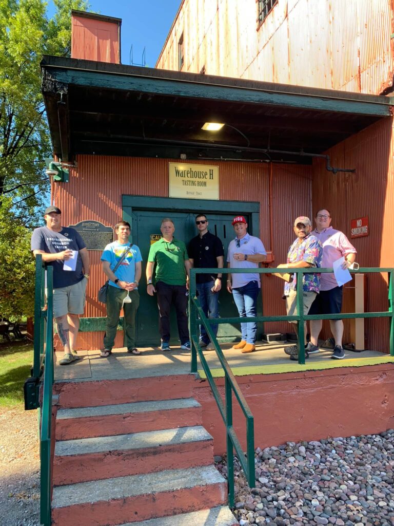 Seven men stand on the porch of a red corrugated-metal building labeled “Warehouse H Tasting Room,” outside a green door in bright sunlight.