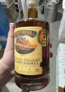Hand holding a “Spirit Rails” Kentucky Straight Bourbon Whiskey bottle (750 ml) with a train illustration on the label, in a warehouse with stacked boxes and a metal keg in the background.
