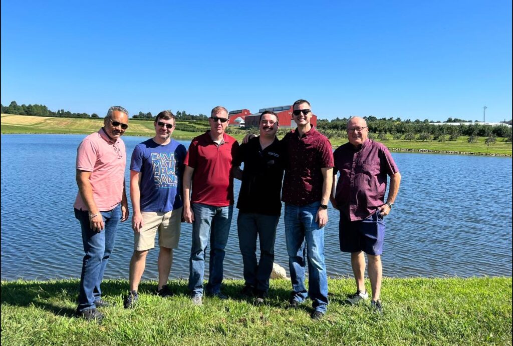 Six men stand together on a grassy bank beside a pond on a sunny day, with rolling farmland and a red barn in the background.