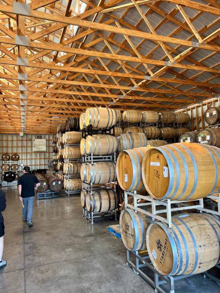 Interior of a large barrel-aging warehouse with wooden roof trusses and rows of oak barrels stacked on metal racks, as two visitors walk down the concrete aisle.