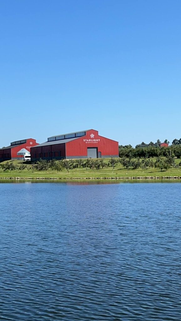 Red Starlight Distillery buildings on a grassy hill across a blue lake under a clear sky.