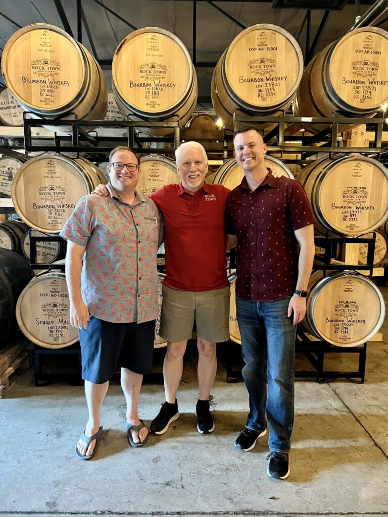 Tyler Shinabery, Phil Brandon, and Alan Myers standing together in front of barrels at Rock Town Distillery, showcasing a friendly atmosphere.