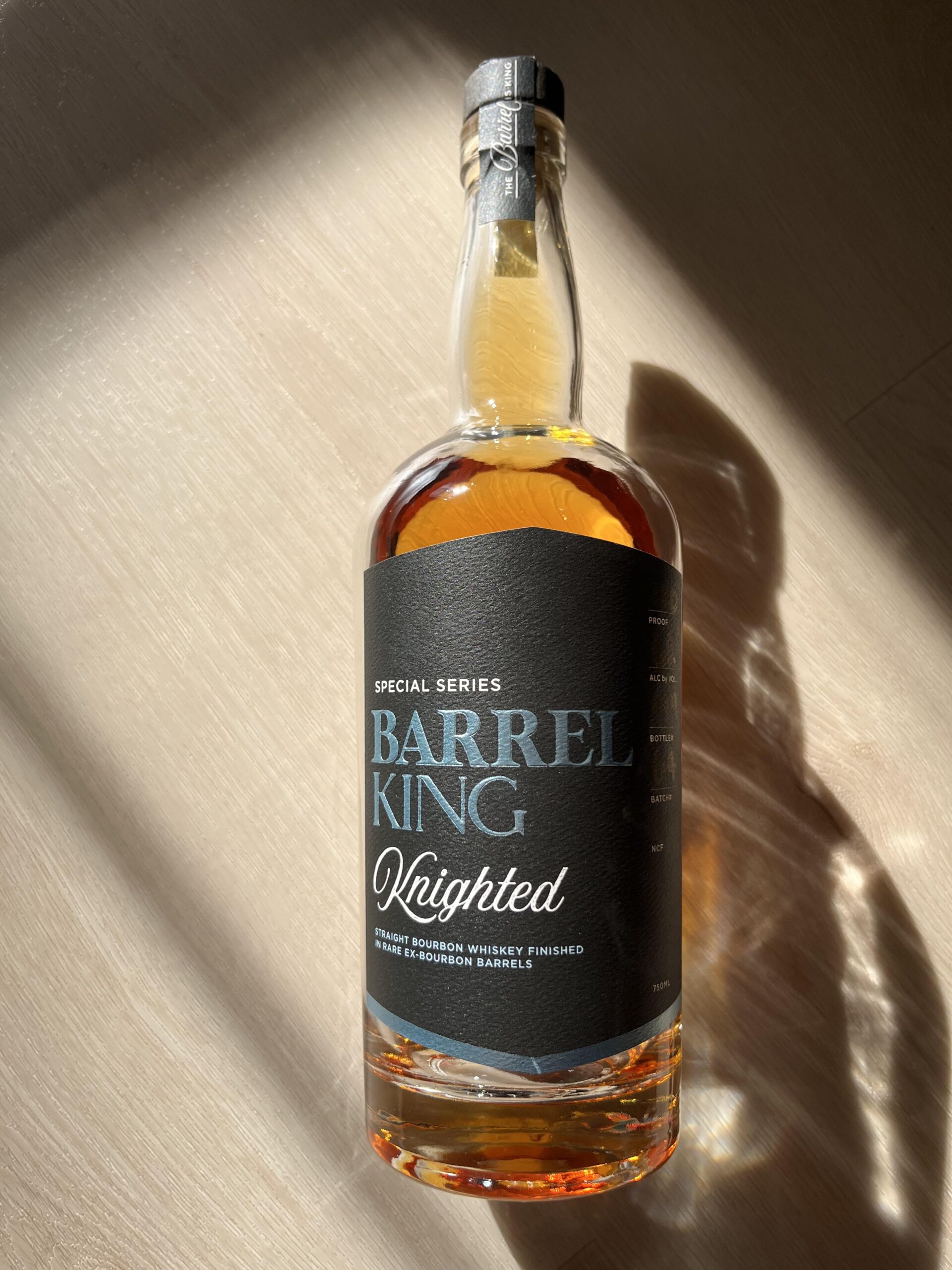 The image shows a bottle of whiskey labeled: - **Special Series** - **BARREL KING** - **Knighted** - **Straight bourbon whiskey finished in rare ex-bourbon barrels** - **750 mL** The bottle is photographed in strong sunlight, casting a sharp shadow on a brushed metal surface.