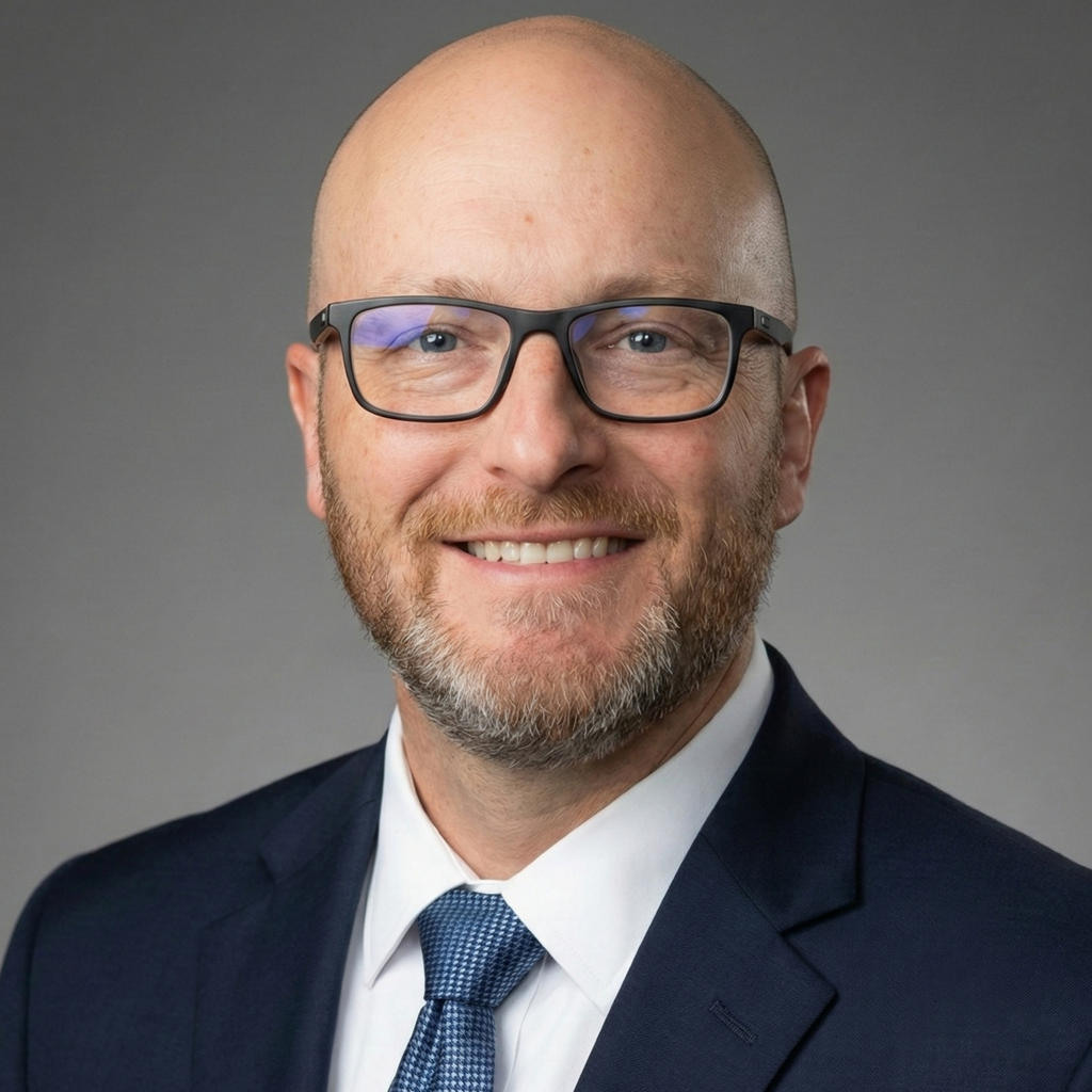 Professional business headshot of a Brian Denham smiling, a bald man with glasses and a short beard, wearing a navy suit, white dress shirt, and blue tie against a neutral gray background.