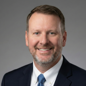 Professional corporate headshot of David Bohannan, a Caucasian smiling middle‑aged man with short brown hair and a neatly trimmed salt‑and‑pepper beard, wearing a navy suit, white dress shirt, and blue patterned tie against a gray studio background.