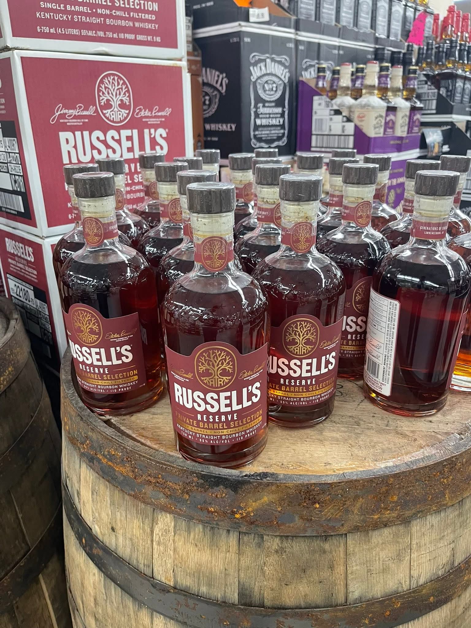Russell’s Reserve Private Barrel Selection bourbon bottles displayed on a wooden barrel in a liquor store, with stacked cases and other whiskey bottles in the background.