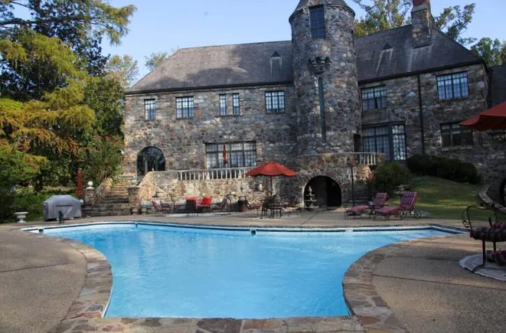 Stone manor house with turret overlooking a backyard swimming pool, patio lounge chairs, and red umbrellas on a sunny day.