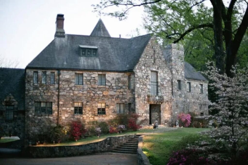 Stone Tudor-style manor house with a steep slate roof and tall chimney, front steps and landscaped garden with spring flowers and mature trees at dusk.