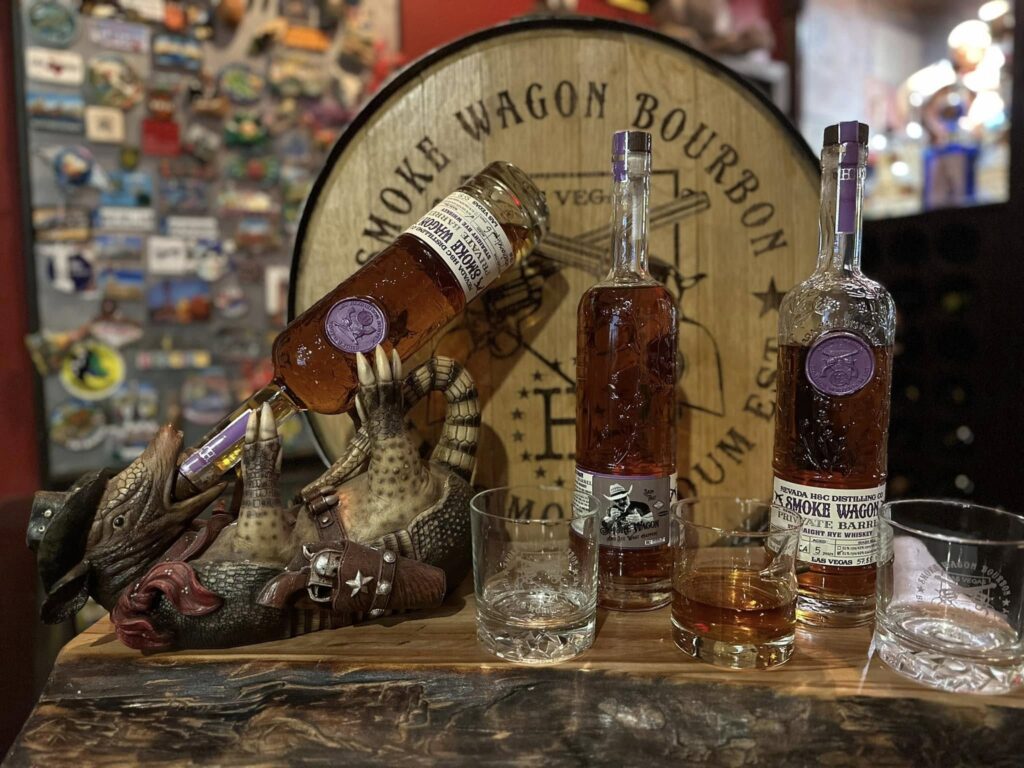 Smoke Wagon Bourbon display with two whiskey bottles, a glass of bourbon and two empty tumblers on a rustic wooden bar, with a playful cowboy armadillo figurine holding a bottle in front of a Smoke Wagon Bourbon barrel head sign.