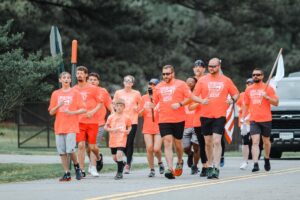 Runners on the street all dressing in orange matching shirts. One person is carrying a white flag with a vehicle behind them.