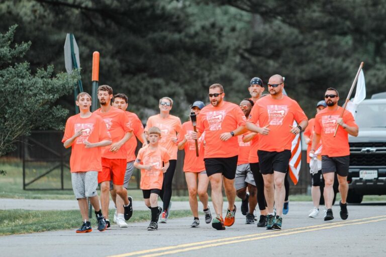Runners on the street all dressing in orange matching shirts. One person is carrying a white flag with a vehicle behind them.
