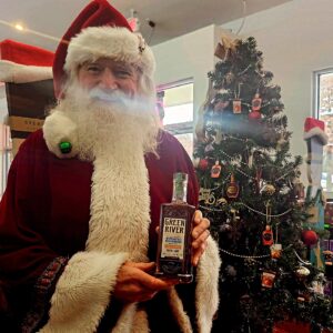 Santa Claus in a red suit and white beard holds a bottle of Green River wheated bourbon in front of a decorated Christmas tree inside a liquor store.