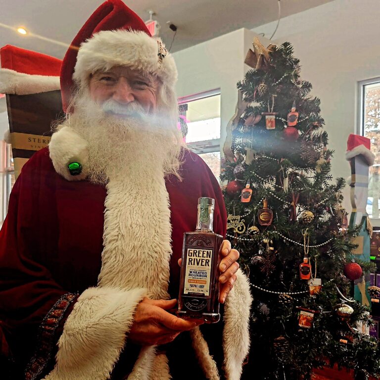 Santa Claus in a red suit and white beard holds a bottle of Green River wheated bourbon in front of a decorated Christmas tree inside a liquor store.