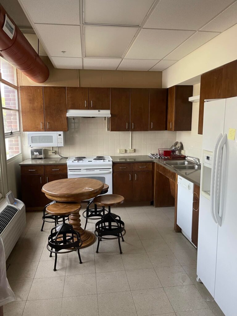 A kitchen space at the Arkansas School for the Deaf featuring wooden cabinets, a stove, microwave, and refrigerator, with a round wooden table and black stools in the center.