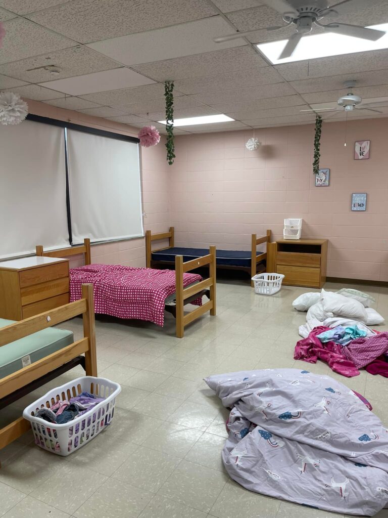A dormitory room at the Arkansas School for the Deaf featuring two bunk beds, scattered bedding, and laundry baskets, with light pink walls and decorations.