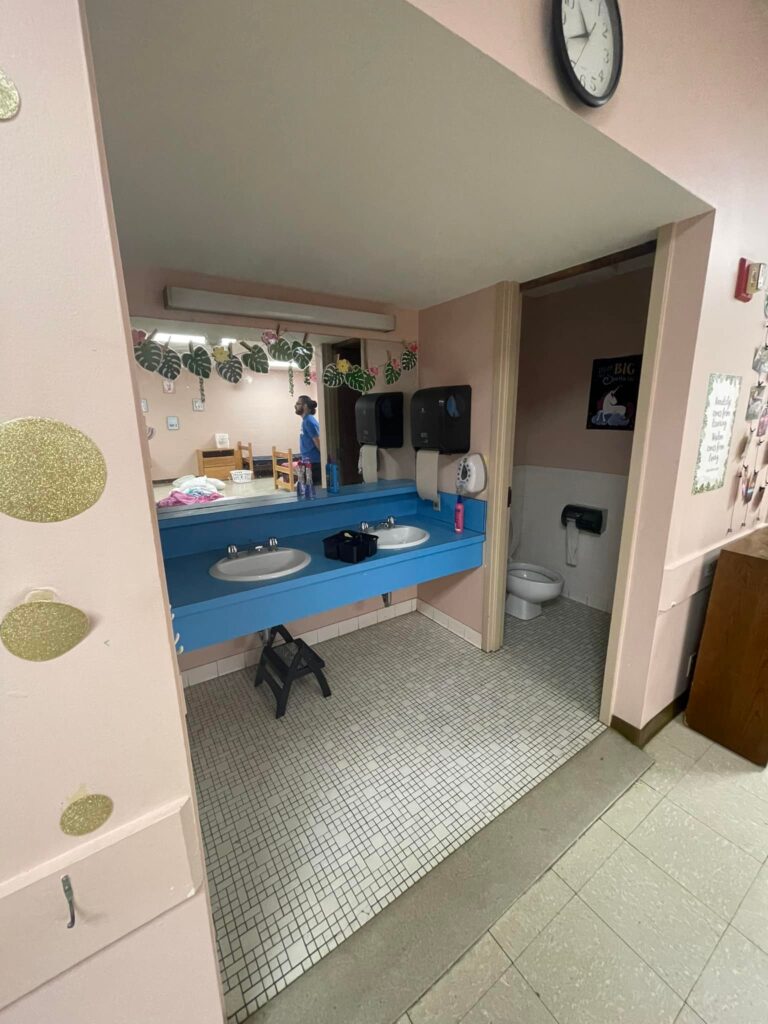An interior view of a restroom at the Arkansas School for the Deaf, featuring a dual sink vanity with blue countertops, transparent mirrors, and soap dispensers, while showing a doorway leading to a toilet area. The walls are adorned with decorative greenery, and the floor is tiled with white and gray patterns.