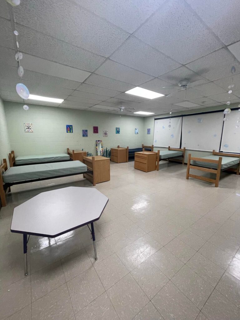 An empty dorm room at the Arkansas School for the Deaf with several unmade beds, wooden furniture, and a small octagonal table, featuring a simple and clean layout.
