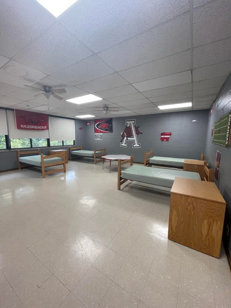 Interior view of a dormitory room at the Arkansas School for the Deaf, featuring empty beds with green mattresses, wooden furniture, and Arkansas Razorbacks decorations on the walls.