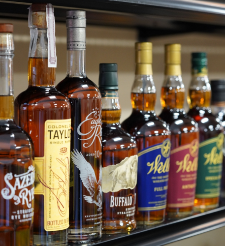 Assorted whiskey and bourbon bottles lined up on a store shelf, including Colonel Taylor, Eagle Rare, and Buffalo Trace in the foreground.