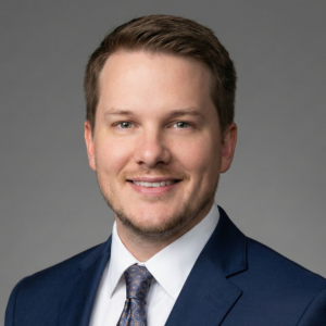 Professional business headshot of Steve Crowe, a Caucasian man with short light-brown hair and light stubble, wearing a navy suit, white dress shirt, and patterned tie against a plain gray background.