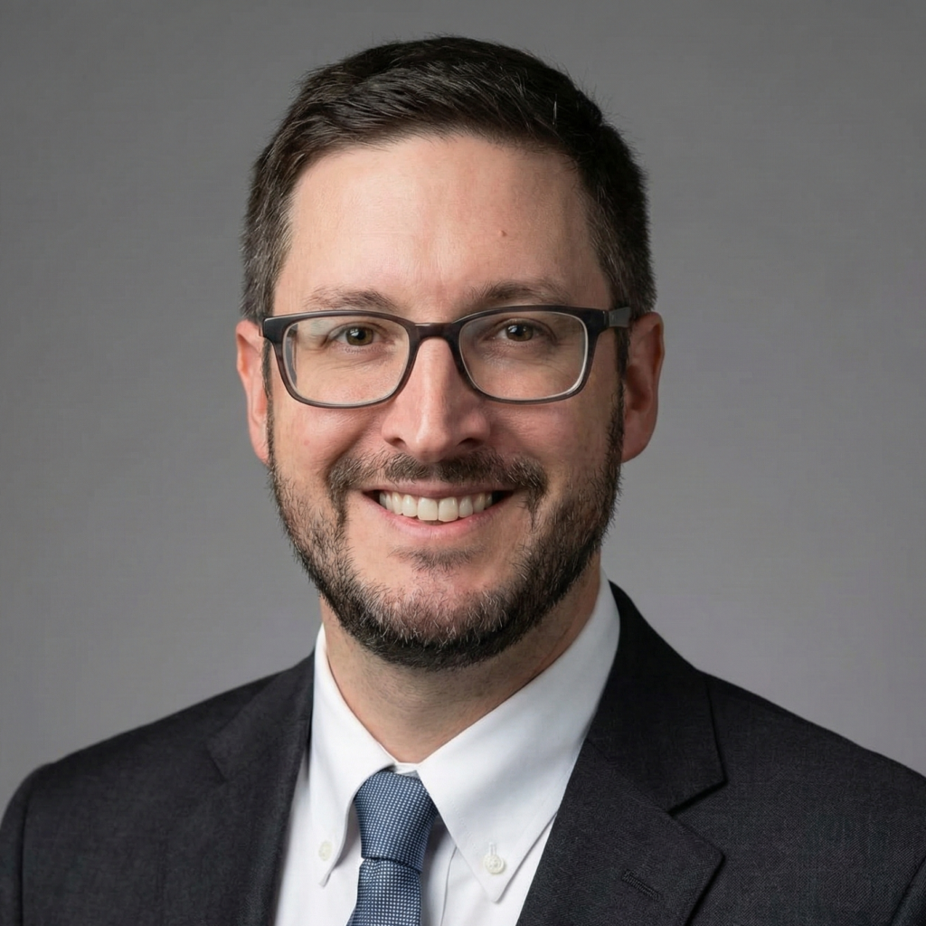 Professional business headshot of Tim Holder, a smiling man wearing rectangular glasses, a dark suit jacket, white dress shirt, and blue tie against a neutral gray background.