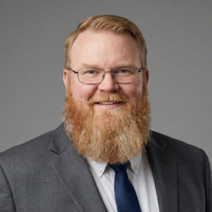 Professional business headshot of Todd Smith, a Caucasian man with short blond hair, rectangular glasses, and a full ginger beard, wearing a dark gray suit, white dress shirt, and navy tie against a neutral gray background.