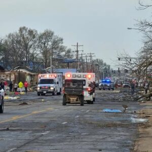 Emergency response vehicles navigating through a storm-damaged street in Arkansas with debris scattered around and people assessing the destruction.
