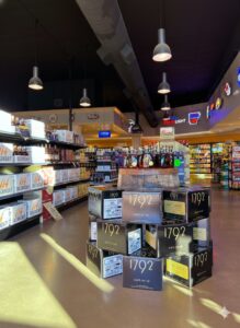 Interior of a liquor store with aisles of beer and spirits; a central display of 1792 bourbon boxes stacked around a wooden barrel, with hanging pendant lights and illuminated brand signs in the background.
