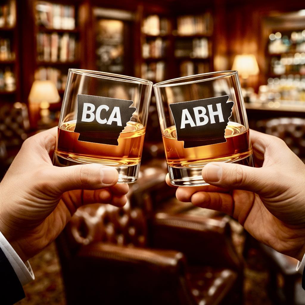 Close-up of two rocks glasses with amber bourbon, each featuring an Arkansas-shaped logo—one says “BCA” for Bourbon Club of Arkansas and the other says “ABH” for Arkansas Barrel Heads—raised in a toast against a warmly lit bourbon bar background.