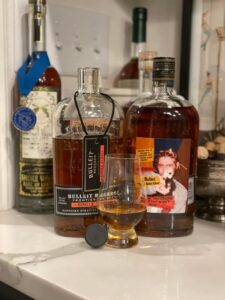 A close-up shot of two bottles of amber bourbon whiskey and a filled tasting glass on a countertop. On the left is a bottle of Bulleit Bourbon Frontier Whiskey Single Barrel with a black label and a neck tag. On the right is a larger bottle labeled Bulleit The Rebel Edition featuring a colorful graphic with a person holding a dog. In front of the bottles is a short whiskey glass with amber liquid. Behind them are other bottles on a shelf slightly out of focus.