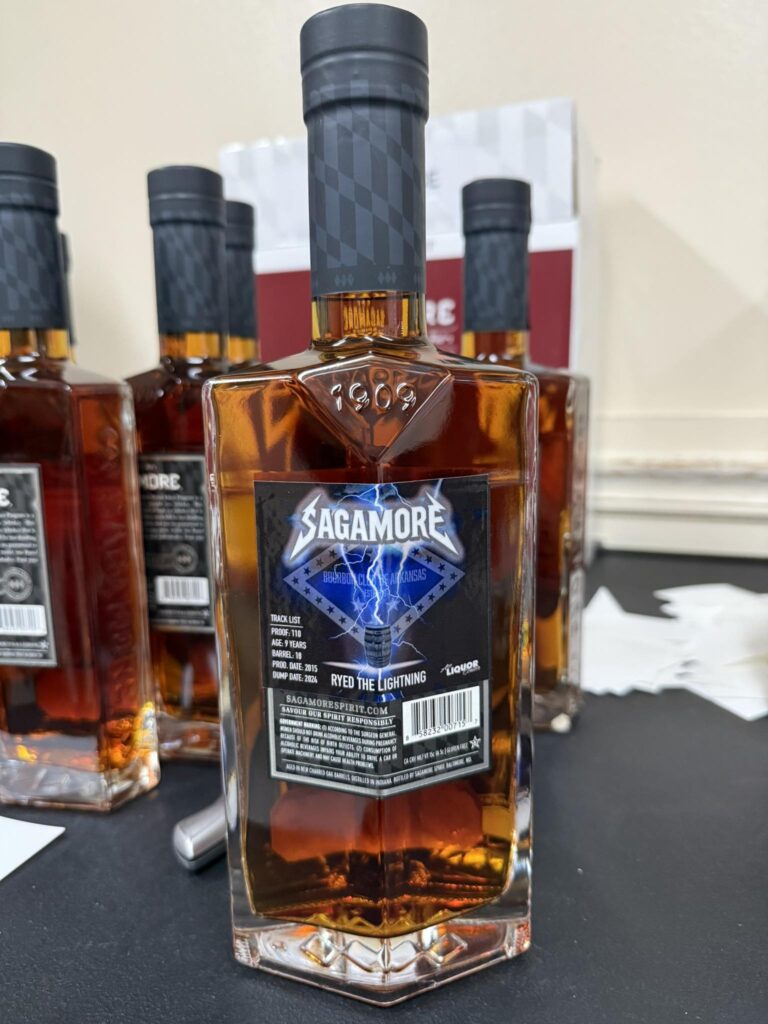 Sagamore Spirit “Ryed the Lightning” rye whiskey bottle (amber liquid) shown close-up on a table, with other Sagamore bottles blurred in the background.