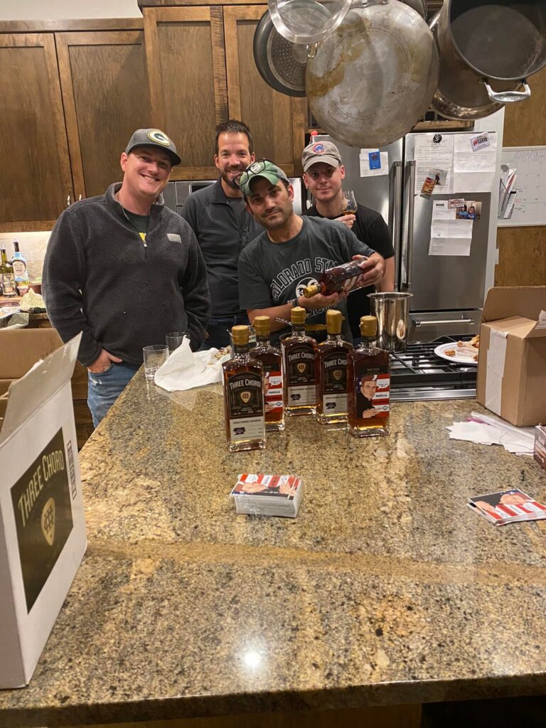 Four men posing behind a granite island. One of them is holding and pouring a bottle of whiskey into a metal cup/jigger. On the counter are several bottles labeled “THREE CHORD” (and a big “THREE CHORD” box on the left), plus a couple of small stacks of cards/packaging. In the background there’s a stainless fridge with papers/magnets and pots/pans hanging overhead.
