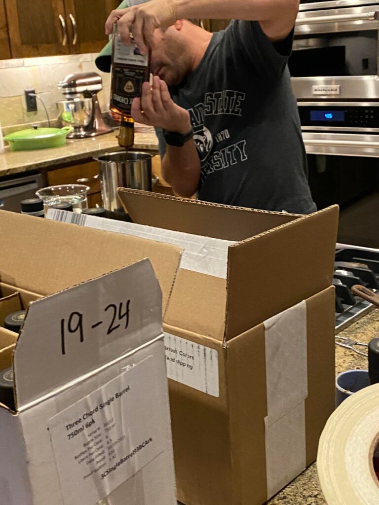 Gary Penna in a home kitchen pours bourbon from a bottle into a stainless-steel flask while packing liquor bottles in cardboard shipping boxes.