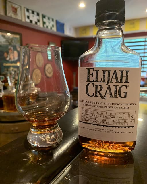 Close-up of an Elijah Craig Kentucky Straight Bourbon Whiskey Private Barrel Program Sample bottle on a dark bar counter, next to a Glencairn tasting glass holding a small pour of amber bourbon; the bottle label reads “ELIJAH CRAIG” with details including barrel number and proof, while a warmly lit whiskey bar or tasting room with framed wall decor and blurred background bottles appears behind, highlighting bourbon tasting, whiskey sampling, and premium Kentucky bourbon.