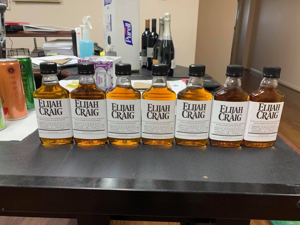 Seven Elijah Craig Kentucky Straight Bourbon Whiskey private barrel program sample bottles lined up side by side on a dark countertop, each with a black cap and white ‘Elijah Craig’ label, showing different amber-gold bourbon shades from light honey to deeper copper. The photo appears to be taken in an office or tasting-room setting; behind the bottles are papers and a desk organizer, a spray bottle and a Purell hand-sanitizer dispenser, plus several unopened wine bottles. The image highlights a bourbon tasting lineup and comparison of multiple Elijah Craig barrel samples in small-format bottles.